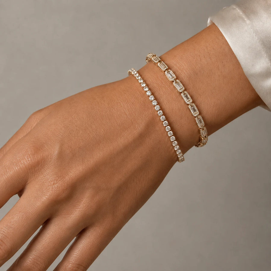 Two delicate tennis-style bracelets on a person's wrist, one in yellow gold with small clear stones and the other in white gold or platinum with larger baguette-cut clear stones, shown against a soft gray background.